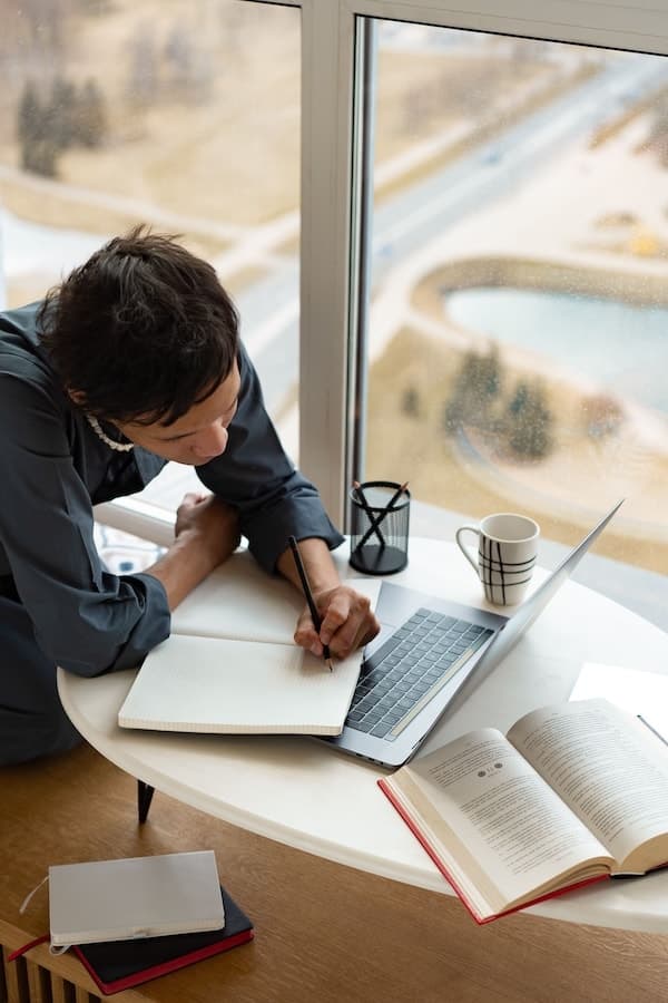 Person schreibt zwischen einem Laptop und mehreren geöffneten Büchern in ein Notizbuch