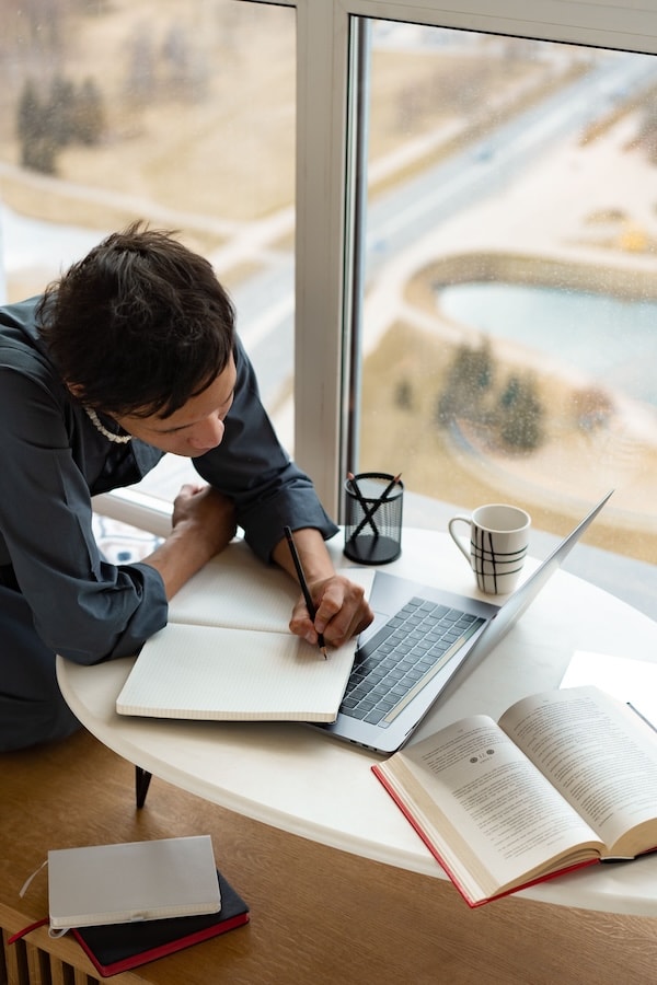 Person schreibt zwischen einem Laptop und mehreren geöffneten Büchern in ein Notizbuch
